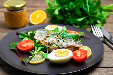 Herring on chewy bread with boiled egg, tomatoes and lemon. Smorobrod with fish and microgrin, on a wooden background. Salad with pickled herring on a black plate.  Scandinavian food.