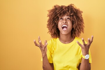 Young hispanic woman with curly hair standing over yellow background crazy and mad shouting and yelling with aggressive expression and arms raised. frustration concept.