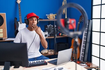 Young hispanic man playing piano recording with smartphone covering mouth with hand, shocked and...