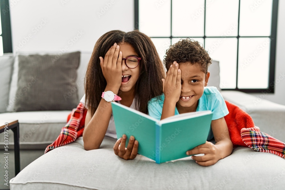 Poster two siblings lying on the sofa reading a book covering one eye with hand, confident smile on face an