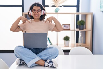 Fototapeta na wymiar Young hispanic woman using laptop sitting on the table wearing headphones looking confident with smile on face, pointing oneself with fingers proud and happy.
