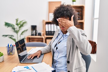 Black woman with curly hair wearing call center agent headset at the office covering eyes with hand, looking serious and sad. sightless, hiding and rejection concept