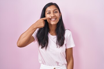 Obraz na płótnie Canvas Young hispanic woman standing over pink background pointing with hand finger to face and nose, smiling cheerful. beauty concept