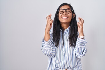 Young hispanic woman wearing glasses gesturing finger crossed smiling with hope and eyes closed. luck and superstitious concept.