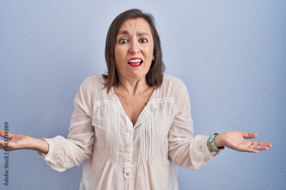 Sticker middle age hispanic woman standing over blue background clueless and confused with open arms, no ide