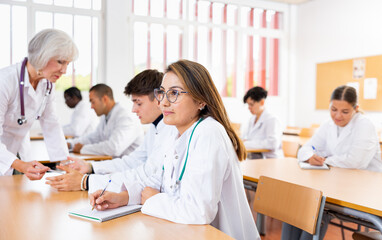 Interested young latin american woman in white coat listening to lecture and taking notes in classroom during professional medical training..