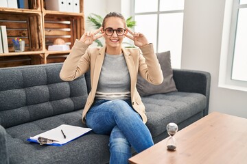 Fototapeta premium Young woman working at consultation office doing peace symbol with fingers over face, smiling cheerful showing victory