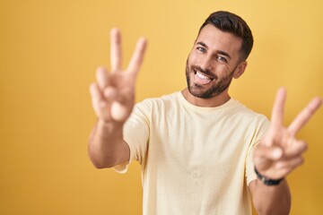 Handsome hispanic man standing over yellow background smiling with tongue out showing fingers of...