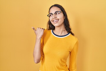 Young brazilian woman wearing glasses over yellow background smiling with happy face looking and pointing to the side with thumb up.