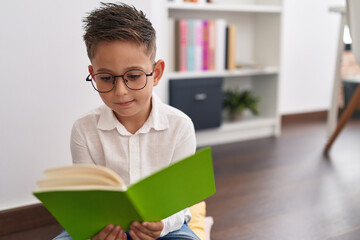 Adorable hispanic boy student smiling confident reading book at library school