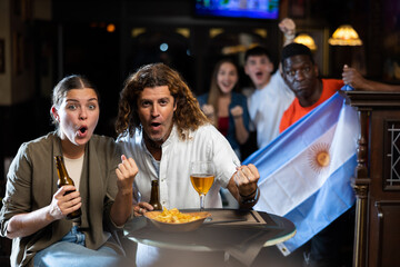 Expressive young adult couple sitting at table in sports bar, drinking beer with snacks and emotionally rooting for favorite Argentinean team while watching football match