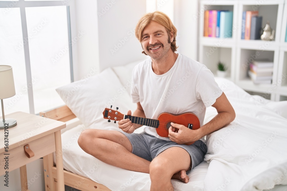 Poster Young blond man playing ukulele sitting on bed at bedroom