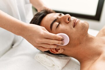 Fototapeta na wymiar Young hispanic man relaxed having facial treatment cleaning face with sponge at beauty center