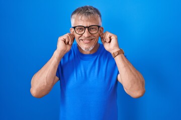 Hispanic man with grey hair standing over blue background smiling pulling ears with fingers, funny...