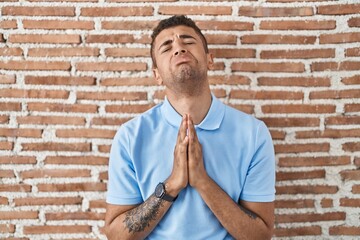 Brazilian young man standing over brick wall begging and praying with hands together with hope...