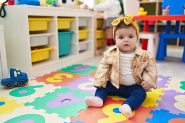 Adorable blonde toddler sitting on floor with serious expression at kindergarten