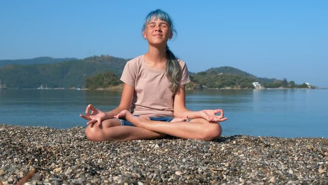 Teen with zen sea practicing. A happy smiling girl sits in lotus position the the pebbles. A concept of sea yoga and relaxation time during summer.