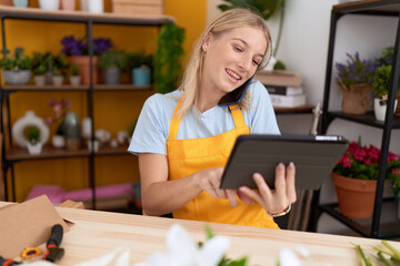 Young blonde woman florist talking on smartphone using touchpad at flower shop