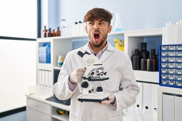 Arab man with beard working at scientist laboratory holding microscope angry and mad screaming...