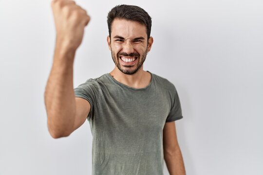 Young hispanic man with beard wearing casual t shirt over white background angry and mad raising fist frustrated and furious while shouting with anger. rage and aggressive concept.