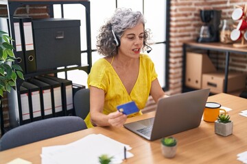 Middle age grey-haired woman call center agent holding credit card working at office