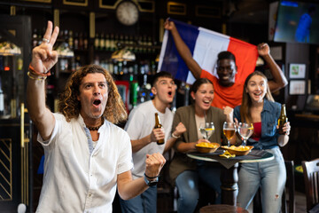 Expressive adult French man cheering for favorite football team while watching game in modern sports bar with group of fans in background