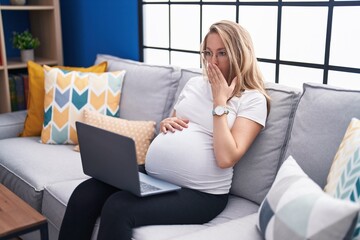 Young pregnant woman sitting on the sofa at home using laptop covering mouth with hand, shocked and...