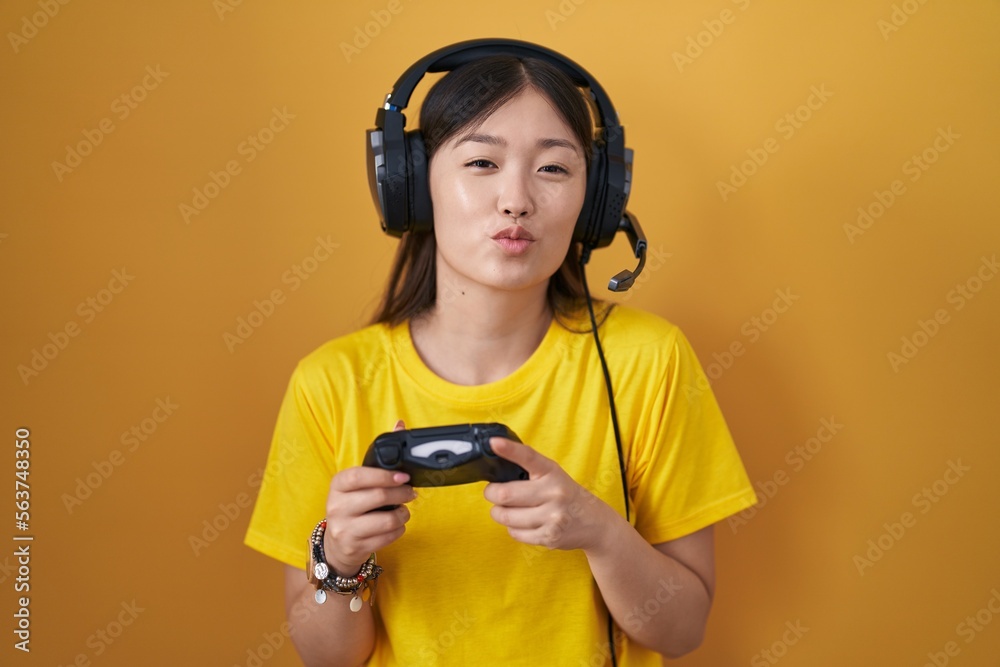 Canvas Prints Chinese young woman playing video game holding controller looking at the camera blowing a kiss on air being lovely and sexy. love expression.