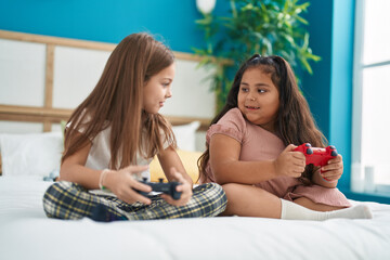 Two kids playing video game sitting on bed at bedroom