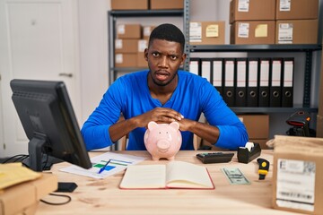 African american man working at small business ecommerce with piggy bank in shock face, looking skeptical and sarcastic, surprised with open mouth