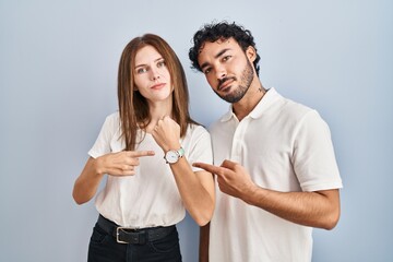 Young couple wearing casual clothes standing together in hurry pointing to watch time, impatience, looking at the camera with relaxed expression