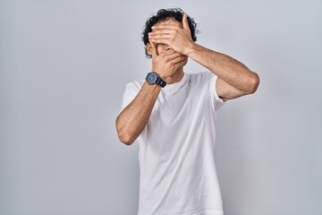 Hispanic man standing over isolated background covering eyes and mouth with hands, surprised and shocked. hiding emotion