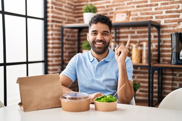 Hispanic man with beard eating delivery salad smiling happy pointing with hand and finger to the side