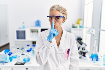 Middle age blonde woman working at scientist laboratory asking to be quiet with finger on lips....