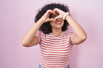 Young middle east woman standing over pink background doing heart shape with hand and fingers smiling looking through sign