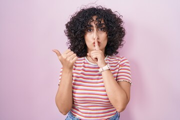 Young middle east woman standing over pink background asking to be quiet with finger on lips pointing with hand to the side. silence and secret concept.