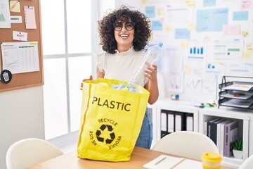 Young middle east woman holding recycling bag with plastic bottles at the office smiling and...