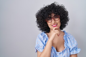 Young brunette woman with curly hair wearing glasses over isolated background looking confident at the camera with smile with crossed arms and hand raised on chin. thinking positive.