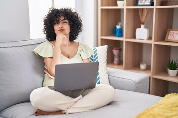 Young brunette woman with curly hair using laptop sitting on the sofa at home looking confident at the camera with smile with crossed arms and hand raised on chin. thinking positive.