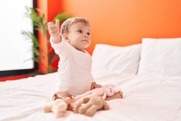 Adorable blond toddler holding doll sitting on bed at bedroom