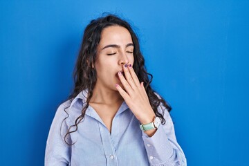 Young brunette woman standing over blue background bored yawning tired covering mouth with hand. restless and sleepiness.