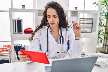 Young hispanic woman wearing doctor uniform using touchpad at clinic