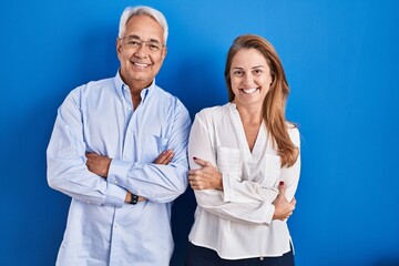 Middle age hispanic couple standing over blue background happy face smiling with crossed arms...