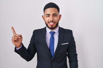Young hispanic man wearing business suit and tie with a big smile on face, pointing with hand finger to the side looking at the camera.