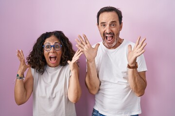 Middle age hispanic couple together over pink background celebrating crazy and amazed for success with arms raised and open eyes screaming excited. winner concept