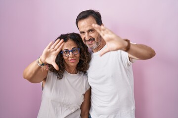 Middle age hispanic couple together over pink background doing frame using hands palms and fingers, camera perspective