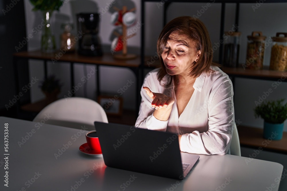 Poster Middle age hispanic woman using laptop at home at night looking at the camera blowing a kiss with hand on air being lovely and sexy. love expression.