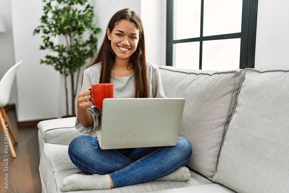 Poster Young latin woman using laptop and drinking coffee sitting on sofa at home