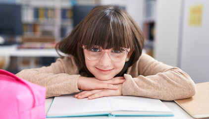 Adorable hispanic girl student sitting on table leaning on book at classroom