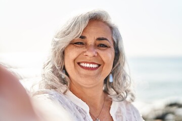 Middle age woman smiling confident making selfie by the camera at seaside